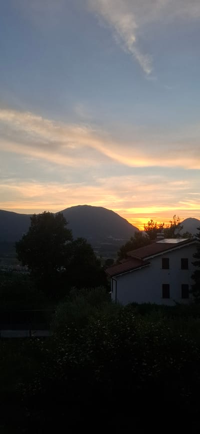 The image depicts a twilight scene with a mountainous landscape under a pastel-colored sky. A house with a red-tiled roof is visible in the foreground, partially silhouetted against the sunset. The house has several dark windows and a visible chimney. Trees and dense foliage are present in front of and around the house, creating dark silhouettes. In the distance, dark mountain ranges are visible, with the setting sun casting a warm glow on the horizon and illuminating the clouds. The overall impression is one of a rural or semi-rural setting at dusk. The specific location in Camerino, Italy, cannot be determined from the visual information alone, as there are no identifying landmarks or features specific to that city.