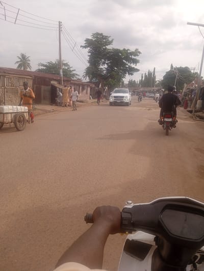 The image depicts a street scene in Tundun Wada South, Nigeria, under an overcast sky, suggesting it is either early morning or late afternoon. The foreground is dominated by the perspective of someone riding a scooter, with their hand gripping the handlebar.
In the middle ground, the street is lined with buildings, some appearing to be shops or homes with corrugated iron roofs. Several people are present. On the left, a man in an orange shirt stands next to a cart with white containers. Further down the street, a group of people in traditional clothing are gathered, possibly socializing or engaged in business.
A white SUV is driving down the road, and several motorcycles are also visible, some with riders and passengers. One motorcycle, in the right middle ground, has a rider wearing a dark shirt and a helmet. The road surface is paved with asphalt.
In the background, trees and more buildings can be seen, and utility poles with electrical wires stretch across the sky. The overall impression is that of a busy, everyday street scene in a Nigerian community. No prominent text is visible in the image, aside from a license plate on the motorcycle in the right middle ground, which reads "KG 4510".