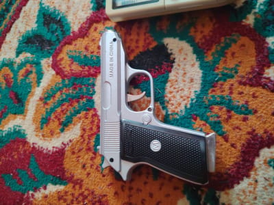 The image shows a toy gun with the inscription "MADE IN CHINA" on the side, resting on a colorful, patterned carpet. Part of a light-colored electronic device is visible at the top edge of the frame, also resting on the carpet. The carpet features a swirling design of red, yellow, green, and black. The setting is indoors, with no visible windows or external environment. There are no people or actions depicted in the image. The image does not provide any information that would allow for the determination of its location in Khujand, Tajikistan, or any other specific city.