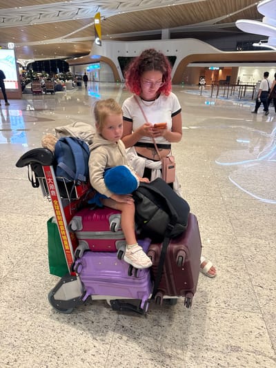 In an airport terminal, likely Baiyun District, China, a young girl with blonde hair sits on a luggage cart laden with several suitcases. She is holding a blue stuffed toy and looking down with a solemn expression. A woman with vibrant pink and red curly hair, wearing glasses and a white t-shirt, stands behind the girl, engrossed in her smartphone. The luggage cart is filled with various bags and suitcases, including a pink suitcase, a purple suitcase, and a dark red suitcase. A green shopping bag hangs from the side of the cart. The floor is a polished speckled tile. In the background, other travelers can be seen walking and sitting, and the airport's modern architecture with illuminated ceiling panels and decorative elements is visible.