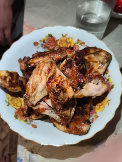 A close-up shot shows a plate of cooked chicken wings. The wings are golden brown and appear to be grilled or roasted, with some darker, charred areas. They are arranged in a pile on a white plate with a floral pattern around the rim. The plate is positioned on a surface that appears to be a table. In the background, a glass of clear liquid is visible, along with parts of what looks like a person's arm and torso. The lighting suggests an indoor setting, possibly a home. The image is likely taken in Il'mino, Russia, based on the prompt, but there are no specific visual cues in the image to confirm this location.
