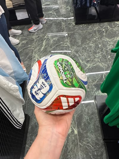 A fair-skinned hand holds a multi-colored soccer ball in a retail environment in Sunrise, United States of America. The ball features white panels with sections of blue, red, and green, displaying the "FIFA" logo (depicting a hand holding a trophy) and text including "TRIONDA" and "REPLIQUE DE BALLON OFFICIEL." The surrounding floor is a polished, dark green, mottled marble-patterned tile with thin white reflective strips.

In the mid-ground, a person's lower legs are visible, clad in black pants and white and grey Nike sneakers featuring a prominent orange stripe on the toe area. This individual stands next to a black retail display unit holding neatly folded white and light grey textiles. Further to the left, another person's legs are partially visible, wearing blue jeans and white sneakers with thick soles. A white garment featuring three black stripes runs vertically near the left edge, and a light blue and white striped fabric is draped. On the right, a vibrant green garment, likely a shirt or jersey, hangs partially visible next to a black display fixture.