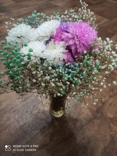 A floral arrangement sits in a glass vase on a wooden surface. The arrangement consists of white chrysanthemums, a purple chrysanthemum, and various small, colorful filler flowers, including green and light blue baby's breath. The background is a brown wooden floor with visible grain. The image is captured indoors, and there is a watermark in the lower left corner indicating "REDMI NOTE 8 PRO AI QUAD CAMERA." There is no indication of the city of Kugesi, Russia, in the image.