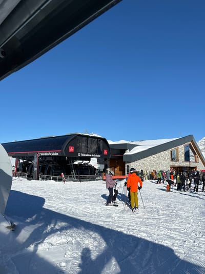 The image depicts a bustling winter scene at a ski resort under a bright, clear blue sky, indicative of excellent weather conditions. The setting is identifiable as Val-d'Isère, France, through the prominent "Télécabine de Solaise" text visible on the black gondola lift cabins.
In the midground, the modern Télécabine de Solaise station is a focal point, with skiers gathering in the snow-covered area around its entrance. Two individuals are notably present on skis near the lift: one wearing a vivid orange jacket and yellow pants, and another in a light purple jacket with black pants. They appear to be preparing to use the lift or have just exited.
To the right, a multi-story building constructed with stone and featuring a grey roof stands amidst the snowy landscape. This structure, likely a mountain restaurant or lodge, is surrounded by numerous skiers and snowboarders, suggesting a popular gathering spot. Their colorful winter clothing adds vibrancy to the snowy environment.
The ground is covered in groomed snow, showing patterns from snow-grooming machines and various ski tracks. Long shadows are cast across the snow, indicating that it is not high noon but likely mid-morning or mid-afternoon, emphasizing the sunny conditions. A dark, angular structure partially frames the scene from the left, possibly part of the lift station or an adjacent building. The overall atmosphere is lively and active, typical of a busy ski day.