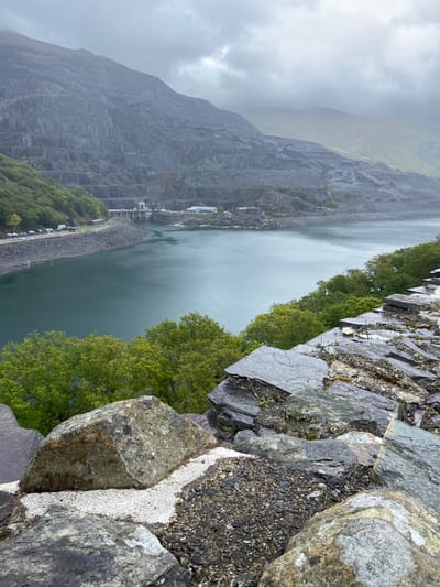 The image depicts a landscape in Llanberis, United Kingdom. A large body of water fills the center of the frame, flanked by a steep, grey, rocky mountain on the left. At the water's edge, there's a road with a row of parked cars alongside green trees. Across the water, more trees line the edge, contrasting with the rocky terrain near the top of the frame. The foreground features rocky ground with larger boulders. The sky is overcast, contributing to a subdued, greyish atmosphere.