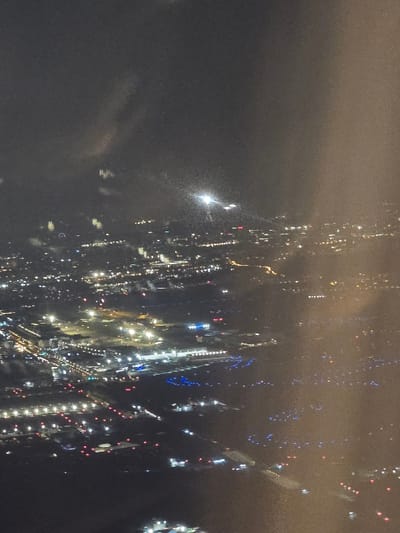 The image displays an elevated, oblique aerial view of a nocturnal landscape in Roissy-en-France, France, consistent with the area around Charles de Gaulle Airport (CDG). The scene is illuminated by extensive artificial lighting under dark, hazy or cloudy atmospheric conditions, with notable light diffusion and reflections present on what appears to be an aircraft window, particularly in the upper and right portions.
The mid-ground and foreground reveal a composite of urban and airport infrastructure. To the left, a dense cluster of white, yellow, and red lights delineates roads and buildings, indicative of a populated urban area. The central and right sections distinctly feature the airport environment, characterized by precise patterns of blue lights marking taxiways and runways, alongside numerous white and red lights indicating various airport facilities, aprons, and guidance systems. Brighter, concentrated clusters of white and yellow lights suggest large structures like terminal buildings or operational centers within the airport complex. A prominent, diffuse bright light source is visible in the upper center, possibly an external ground light or reflection. No discernible human figures or specific aircraft are present. The scene implies ongoing activity within both the urban and airport landscapes, with vehicular movement on illuminated thoroughfares and airport operational zones.
