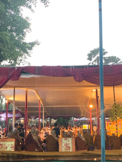 The scene is a gathering of people under a large tent in Mungkid, Indonesia, located at coordinates -7.605, 110.23. The tent is made of white and red fabric, and there are numerous support poles. There is a group of people in orange robes sitting in the front row, facing the camera. Behind them, there are other people seated, mostly in casual clothes. In the background, more people are gathered, and some appear to be dancing or performing. The scene appears to be a gathering or celebration, possibly a religious event. The weather appears to be rainy, as raindrops can be seen falling from the tent's fabric. The sky is overcast, with a white background. There are trees and lush green foliage surrounding the tent. The photo is taken from a low angle, looking up at the scene. The camera is focused on the group of people in orange robes, with the rest of the scene slightly out of focus. The image is slightly dark, but still clear enough to make out the details. The photo was taken at dusk, with the lighting suggesting that the event is taking place at night.