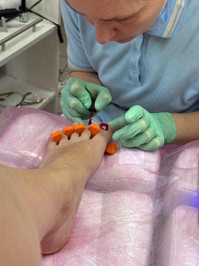 A close-up shot shows a person's foot being given a pedicure. The toes are separated by orange foam toe separators. A person wearing light blue medical scrubs and green gloves is applying dark red nail polish to the big toe. The foot rests on a pink paper-like surface. In the background, there are shelves with medical instruments and what appears to be part of a UV lamp for drying nail polish. This scene is taking place in a nail salon in Murom, Russia.