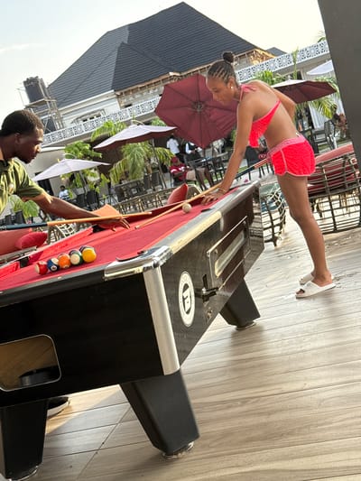 The image captures a moment in Rubuchi, Nigeria, featuring a woman in a red bikini and red crochet shorts playing pool on an outdoor table. She is bending over the table, cue in hand, about to strike the cue ball. A man stands on the opposite side of the table, observing. The pool table is positioned on a wooden deck, and the backdrop includes outdoor seating areas with maroon umbrellas. There are several unidentified individuals seated in the background. The weather appears sunny.
