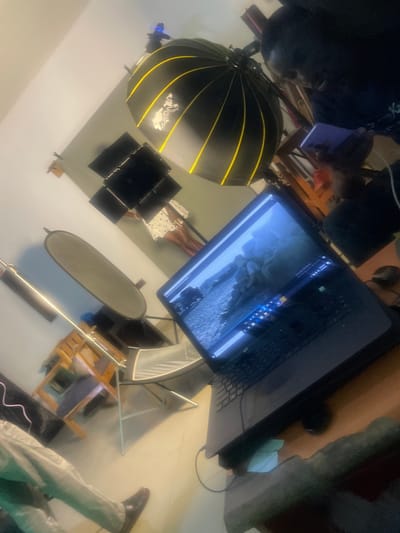 The image depicts an indoor scene that appears to be a photography studio or a similar creative space, possibly located in South Ropp, Nigeria. The perspective is tilted, giving a sense of depth. In the foreground, a laptop is open, displaying a picture of rocks and water. To the left of the laptop, there's a large, parabolic-shaped light modifier with yellow stripes. Behind the laptop, a person is visible, wearing dark clothing and looking down at a smartphone. Further back, a person in a white polka-dot dress is standing near a set of black rectangular light panels. The room contains various studio equipment, including light stands and what looks like a collapsible backdrop or reflector. Furniture, including chairs and what might be a wooden easel or shelving unit, is also present. The overall lighting suggests artificial illumination, consistent with a studio environment. The floor is tiled.