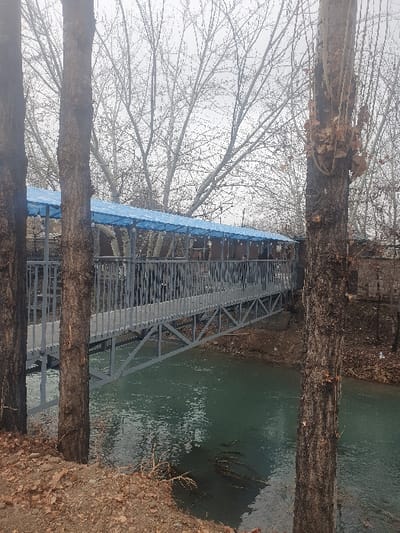A pedestrian bridge, constructed of grey metal trusses and vertical slat railings, spans a light green body of water in Uchqo'rg'on, Uzbekistan. The bridge superstructure is covered by a continuous light blue tarp canopy. Deciduous trees, primarily bare of leaves, line both banks and form the background beneath an overcast, grey sky. Three large, dark-barked tree trunks are prominent in the foreground, partially framing the bridge; the trunk on the far right retains clusters of dry brown leaves. The foreground ground consists of brown soil, dry leaves, and minimal dry vegetation. No individuals or active subjects are present on the bridge or within the visible scene.