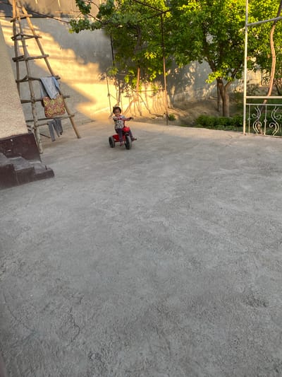 The scene, likely in Akhtachi, Uzbekistan, shows a concrete yard on a sunny day. A young child is riding a red tricycle in the middle of the yard. The yard is enclosed by walls, with a fence visible in the background. A tree and some wooden structures (possibly a clothes rack) are also present in the yard. Clothes are hanging on the rack.