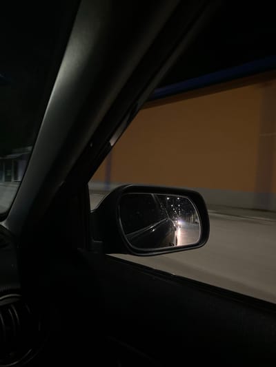The image captures a nighttime view from inside a car, looking out the passenger side window and into the side mirror. The car appears to be in motion on a road, indicated by the blurry streaks of light in the distance. The side mirror reflects the road behind the car, showing another vehicle with its headlights on, illuminating the asphalt. The environment outside the car consists of a building with an orange-colored facade, partially obscured by the car's window frame. The sky is dark, suggesting it is nighttime. There is no specific information to identify the location as Yasen, Bulgaria.