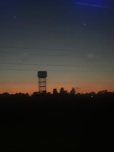 The image depicts a power line tower against a twilight sky. The sky transitions from a pale blue on the left to a warm, orange hue near the horizon, before deepening into blackness at the right. Silhouetted trees line the horizon, obscuring the ground. The power lines are visible as thin, dark lines stretching horizontally across the frame. A small, dark, rectangular structure is attached to the tower's central support. A faint, bright point of light, possibly a star or distant aircraft, is visible in the upper right quadrant of the sky. Another faint blue light is visible in the lower left quadrant. The scene suggests an outdoor setting at dusk or dawn, with the power lines indicating an area with electrical infrastructure. No people or specific activities are depicted, and no text is visible. The location context is Hod HaSharon, Israel.