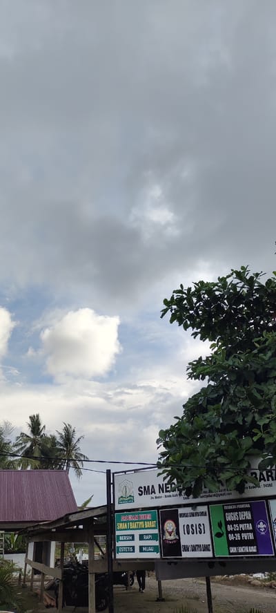 The image captures an outdoor scene in Langnibong, Indonesia, under a cloudy sky. In the foreground, a dense green tree with large leaves occupies the right side of the frame, obscuring part of the sky. To the left, a building with a dark red, corrugated metal roof is visible, with wooden supporting structures in front of it. Several motorcycles are parked under this shelter. Adjacent to the building is a signpost with multiple rectangular panels displaying text and graphics. One panel, with a green background, reads "PROGRAM BERES SMAN 1 BAKTIYA BARAT" and lists qualities like "BERSIH," "RAPI," "ESTETIS," and "HIJAU." Another panel features the text "(OSIS)" and the name "SMAN 1 BAKTIYA BARAT." A third panel announces "GERAKAN PRAMUKA GUGUS DEPAN 04-25 PUTRA 04-26 PUTRI SMAN 1 BAKTIYA BARAT," accompanied by scouting emblems. A larger sign above these smaller panels displays "SMA NEGERI" and some text in smaller font. In the background, palm trees are visible, suggesting a tropical or subtropical environment. The overall impression is a school or community area on a partly overcast day.