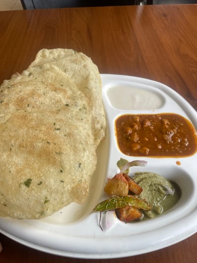 A white disposable plate, containing a meal, rests on a wooden table. The plate has three compartments. The largest compartment, on the left, holds two pieces of puffy, golden-brown fried bread, possibly poori, with visible green herbs sprinkled on them. To the right of the bread, a compartment contains a thick, reddish-brown curry, likely chana masala, with visible chickpeas. The smallest compartment, at the bottom right, holds a dollop of pale green chutney, a few pieces of fried potato or paneer, a sliver of red onion, and a piece of green chili pepper. A creamy white sauce is in the top right compartment. The background shows the texture of a wooden table. The scene is likely from Sidhpur, India, a city known for its culinary heritage.