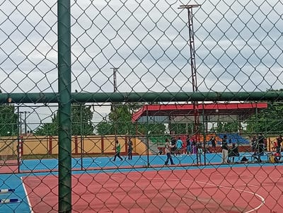The image depicts an outdoor multi-purpose court at the Adeniran Ogunsanya College of Education in Ijanikin, Nigeria, viewed through a chain-link fence. The court is divided into a red section and a blue section. Several individuals are on the court, some walking, some standing. A building with a red roof is visible in the background, behind a low wall. There are tall light poles in the distance. The sky is overcast.