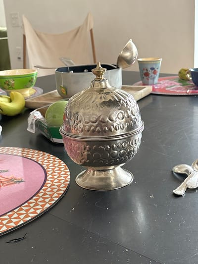 The image shows a silver sugar bowl with a lid, on a black wooden table in a dining room. The bowl is centered in the image. It has a floral design on its surface and a gold knob on its lid. To the left of the sugar bowl is a pink-colored, circular placemat with a white, geometric design around the edge.  The placemat has a floral design in the center. There is a green, ceramic bowl to the left of the placemat with a few green bananas in it. There is a large, silver ladle to the top right of the bowl.  The scene is located in the town of Celerina/Schlarigna, Switzerland. The coordinates 46.514, 9.859 place the scene in the center of the town.  The wooden table is clean except for a few crumbs and a hair to the left of the sugar bowl.  There is a silver fork off to the right of the sugar bowl.