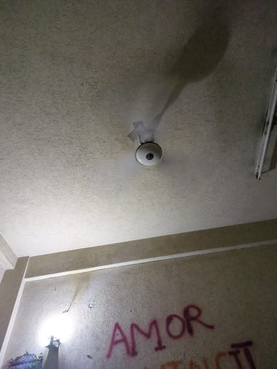 This image shows a textured ceiling in a room, likely in Abbottabad, Pakistan. A ceiling fan is mounted on the left side of the frame, casting a shadow. The fan appears to be white with a circular light fixture. On the right side of the ceiling, graffiti in red spray paint is visible, reading "AMOR". The scene appears to be indoors, with artificial lighting present, suggesting it could be nighttime or a dimly lit interior. The texture of the ceiling is rough and uneven. The overall impression is of a somewhat neglected or vandalized space. No people are visible in the image.
