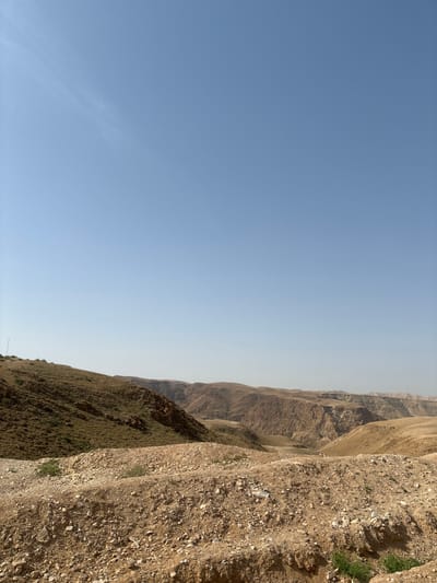 The scene displays an expansive arid, mountainous landscape under a clear, pale blue sky in the Megilot Yam HaMelakh Regional Council, Palestinian Territory. The foreground features a light brown and beige rocky slope covered in loose gravel and small stones, with isolated patches of green scrub vegetation. To the immediate left, a larger hill is present, characterized by patchy olive-green vegetation and visible reddish-brown rock outcrops. A thin, dark, vertical object, possibly an antenna or pole, is faintly discernible on the upper left slope near the horizon. The midground and background consist of successive layers of barren, eroded mountains and deep valleys, presenting a spectrum of tan, brown, and ochre tones, gradually fading into a light atmospheric haze. No individuals, animals, specific actions, or interactions are visible.