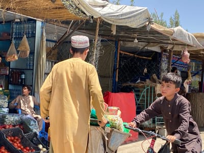 The scene takes place in a marketplace in Dand, Afghanistan (latitude: 31.617, longitude: 65.628). A man in a tan long-sleeved shirt and a red and white skullcap is standing in front of a storefront, facing the left. He is looking off to the side and slightly down, not making eye contact with the viewer. He is the central subject of the image. To his left, a young boy in a beige shirt and dark pants is sitting on a chair and looking to the left of the frame. He is holding his arms folded across his chest. Behind the boy is a stall with a variety of items for sale including tomatoes, onions, and bottled beverages. Further in the background, a man in a brown shirt is standing by a stall.
To the man in the tan shirt's right, another young boy in a long brown shirt and pants is riding a red bicycle towards the right of the frame. He is looking ahead at the road. His right hand is on the handlebars, while his left hand is holding onto the seat. The boys' actions and the man's stance suggest that this is a busy marketplace with people moving and interacting with each other.
The scene is set under a partially covered roof, with the open side facing the street. There are visible thatched straw structures on the roof. The area is sparsely decorated and appears to be in an older state of repair. The presence of the bicycle suggests that this is a popular place to shop and socialize.