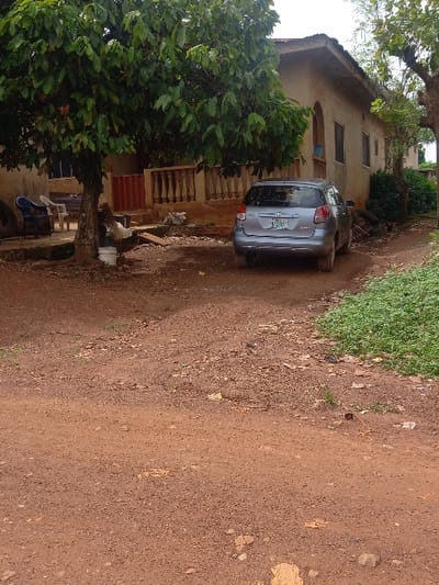 The image shows a silver Toyota Corolla parked in front of a single-story house in Ita Oniyan, Nigeria. The house is made of concrete and has a red-brown facade. There is a tree to the left of the house, with a small, light-colored bucket sitting underneath it. The ground in front of the house is dirt and gravel, with some green grass patches. The car is facing the camera, with a license plate visible on the rear bumper. There are no people visible in the image. The coordinates 7.291 latitude and 5.166 longitude, place this scene in a residential area of Ita Oniyan.