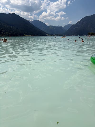 This media file captures a scenic view of a lake or swimming area in Lärchenwiese, Austria. The primary subjects are people enjoying the water and the natural surroundings. In the foreground, several individuals are partially submerged in the light blue, slightly murky water, appearing to be swimming or wading. One person in the lower right is wearing a hat, and another has a tan torso. Further in the water, other people are visible as dark shapes.

The background is dominated by a dramatic mountain landscape. A steep, dark green, tree-covered mountainside rises from the shoreline and extends upwards, meeting a bright blue sky filled with fluffy white clouds. The sun appears to be high in the sky, suggesting it is daytime, likely midday or early afternoon, with bright lighting illuminating the scene. A large, bright green inflatable object is partially visible at the top of the frame, hinting at recreational activities on the water. The water itself exhibits a rippled texture, reflecting the light and creating a dynamic surface. The overall impression is one of a pleasant, active summer day spent outdoors in a beautiful alpine setting. No text is visible in the image.