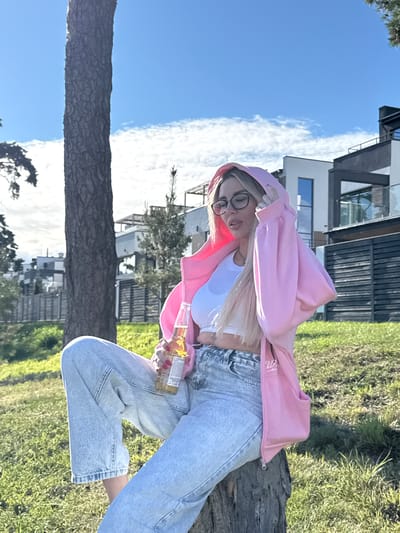 A blonde woman wearing glasses, a white crop top, light blue jeans, and a pink hoodie sits on a tree stump outdoors. She is holding a bottle of beer. The background shows a modern house with dark and white elements, a wooden fence, and green grass. The sky is blue with scattered clouds. The location appears to be a suburban area with residential buildings.