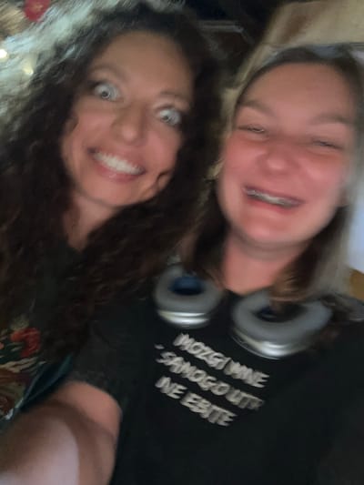 Two women are in a close-up selfie. The woman in the foreground has dark curly hair and is smiling widely. The woman in the background has light brown hair, braces, and headphones around her neck. The background is out of focus but appears to be indoors. The location is not discernible as Bắc Hà, Vietnam from the image. The text on the shirt is unintelligible.