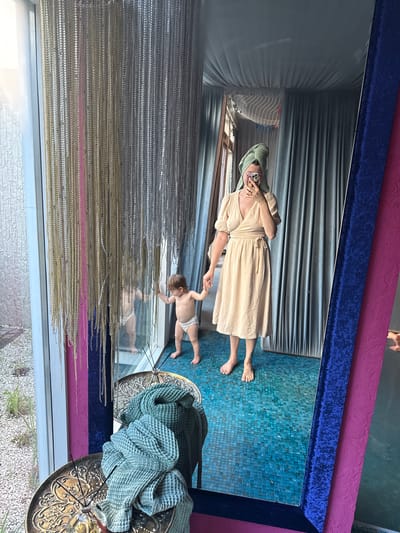 The image shows a reflection in a mirror with a blue and purple frame. A woman wearing a light-colored dress and a head wrap is standing on a blue tiled surface and holding a smartphone to take a picture. A child in a diaper is reaching towards the woman. The reflection shows a gray curtain behind the woman and a draped netting structure. The foreground of the image shows a window with beaded strands and the exterior featuring small pebbles and vegetation. The location of Чёрное, Russia is not discernible from the visual information.