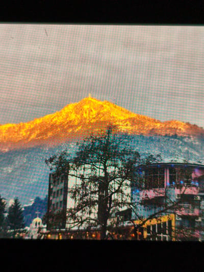 The image depicts a cityscape of Devin, Bulgaria, at dawn or dusk. A mountain, likely part of the Rhodope Mountains, dominates the background, with the summit brightly illuminated by sunlight, appearing golden. The lower slopes are shadowed, suggesting a contrast in lighting conditions.
In the foreground, a large tree with bare branches partially obscures several buildings. To the left, a white church with a golden dome and cross is visible. A taller, more modern building is adjacent to the church, while a pink building with illuminated windows stands to the right. Streetlights glow along the bottom of the frame. The sky is overcast and pale.