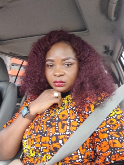 An adult female is depicted in a medium-shot portrait, seated in the front passenger or driver's seat of a vehicle, secured by a grey seatbelt. She has dark skin and voluminous, curly, reddish-brown hair. Her facial features include red lipstick, defined eyebrows with light-colored pigment, and light eye makeup. She is wearing a short-sleeved top with a black background and orange and yellow abstract patterns. A dark brown smartwatch is visible on her left wrist, and a large, light-colored ring adorns the ring finger of her right hand, which is positioned near her chin. Her expression is neutral, and her gaze is directed towards the camera.

The vehicle's interior features a grey headliner with a visible sunroof. Through the window on the left side, a multi-story building with orange walls and dark-framed windows is partially visible in the blurred background. The scene is located in Festac Town, Nigeria.