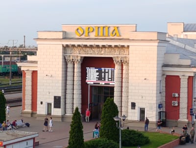 The image depicts the Orsha railway station in Belarus on a clear day, likely in the late afternoon or early evening given the warm lighting. The station building is a prominent architectural feature, characterized by its large, arched entranceway topped with the station's name, "ОРША," in large yellow letters. The facade is made of pale, cream-colored bricks, accented with decorative carvings and large columns flanking the entrance.
Inside the entrance, a digital display board shows train schedules. Several people are visible in the scene. On the concourse in front of the station, a few individuals are waiting or walking. One man in a red shirt stands near the entrance. To the left, near a bench, a small group of people are seated, with luggage nearby, suggesting they are travelers. Further to the left, a man stands alone, looking towards the railway tracks, where trains are visible in the background. On the right side of the station, a few more people are seen in the distance, some with backpacks. The surrounding area features green trees and manicured bushes, with a modern streetlamp visible in the foreground. The overall atmosphere appears calm, with people engaged in typical activities at a train station.