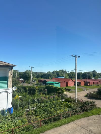 The image depicts a residential area in Rasskazovo, Russia, characterized by a clear blue sky and abundant greenery. In the foreground, a garden plot is visible, filled with various plants, flowers, and two large blue barrels. A rusty metal fence separates the garden from a dirt road and a grassy area. To the right of the garden, a row of red and green garages stands adjacent to a dirt path. In the background, numerous trees form a dense forest, with some residential structures interspersed. Power lines and utility poles are prominent features throughout the scene. On the left side of the image, the corner of a building with a corrugated roof is visible, showing white and green siding. A satellite dish is mounted on the exterior wall of this building, overlooking the garden. The overall impression is that of a rural or semi-rural environment in the summertime.