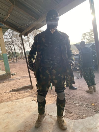 An outdoor scene in Zaki Biyem, Nigeria, depicts two individuals in military-style camouflage attire. In the immediate foreground, a person stands centrally on a light-colored concrete surface, directly facing the viewer. This individual wears a camouflage uniform (green, brown, light-patterned), a dark balaclava or face mask featuring a light-colored skeletal design around the mouth, and dark goggles or sunglasses that reflect bright sunlight. Black gloves with a distinct pattern cover their hands. They are equipped with light-colored tactical knee pads, marked "TSUS" and "KCUNE," over their trousers, and light-colored tactical boots. The person's right hand (viewer's left) holds a dark, elongated object pointed downwards.
In the mid-ground to the right, a second individual in camouflage uniform is visible from the back, wearing a dark tactical vest or backpack and light-colored boots. Behind this person, a dark-colored vehicle, possibly a truck or SUV, appears with an open hood or damaged front, illuminated by a strong, low sun creating lens flare.
The environment features a reddish-brown dirt ground with some scattered debris. To the left, a portion of a building with a green-painted base pillar is visible. An overhead corrugated metal roof structure supported by wooden beams provides shade for the foreground. In the far background, numerous leafless trees stand under a bright, clear sky, characteristic of a dry season or arid climate.