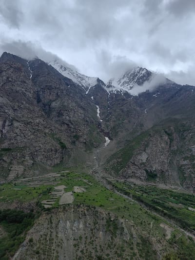 This is a vertical, eye-level, wide shot of a mountain landscape in Gemur, India, under a cloudy sky. In the foreground, a steep, rocky mountainside slopes down to the left, with patches of green vegetation scattered across its surface. In the middle ground, several terraced fields are visible on the green mountainside, some appearing to be cultivated. A narrow, winding path is visible to the far right of the terraced fields. The background features massive, rugged mountains, with snow-capped peaks visible at the very top. Clouds obscure parts of the mountain peaks and drift across the sky, creating a dramatic and somewhat overcast atmosphere. A small stream or ravine can be seen cutting through the mountainside in the middle ground, with patches of snow still lingering in shaded areas. The overall scene depicts a remote and natural mountain environment with evidence of human cultivation in the form of terraced agriculture.