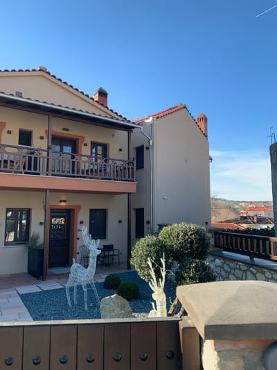 The image captures an outdoor scene in Kastraki, Greece, featuring a picturesque courtyard flanked by traditional buildings under a clear blue sky. Two multi-story buildings, characterized by their light-colored (beige/cream) facades and terracotta-tiled roofs, dominate the left and center of the frame. The building in the foreground has a prominent wooden balcony with intricate railings, and several windows and doors are visible, some with dark frames. A similar building stands slightly behind it. Both structures exhibit features common to Mediterranean architecture, including exposed brick chimneys and simple, clean lines.
The courtyard area in the foreground and center is covered with light grey gravel, interspersed with lighter paved sections. Decorative landscaping includes several round, green bushes and a large, white wireframe deer figure, possibly serving as an outdoor ornament or seasonal decoration. Near one of the doorways of the foreground building, a small patio set consisting of two black chairs and a table is visible. Further right, a partial view of a dark wooden gate or fence post with prominent metal studs frames the edge of the image.
In the upper sky, a faint white contrail indicates the path of an aircraft. Beyond the buildings, a distant hilly landscape with scattered red-roofed houses suggests a village or town built into the terrain. The bright, clear blue sky and the lighting indicate it is daytime, likely a sunny morning or afternoon. There are no people visible, and no discernible text appears in the image. The overall impression is one of a serene and well-maintained outdoor space in a traditional Greek setting.