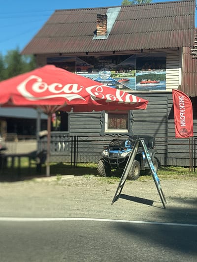 A sunny outdoor scene in Katun', Russia, features a red Coca-Cola umbrella in the foreground, casting a shadow. Behind the umbrella, a dark wooden building displays advertisements for outdoor activities such as jeep tours, quad biking, and raft trips. A blue and black ATV is parked near the building, next to a foldable A-frame sign. To the right of the building, a vertical red flag with Russian text and a logo hangs. The ground is a mix of gravel and grass. The building's roof is made of corrugated material, and a brick chimney is visible. The sky is clear and blue.