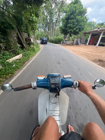 The image, taken from the first-person perspective of a person riding a blue Honda motorcycle, shows a road in Angulugaha Junction, Sri Lanka. The rider's hand is on the right handlebar. The road curves to the left, and a blue vehicle is visible in the distance. Buildings and vegetation line the roadside. The sky is overcast.