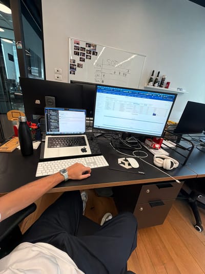 The image depicts a workspace, likely an office, with a desk cluttered with technology and personal items. A person is seated at the desk, using a white keyboard and wearing a silver wristwatch. On the desk, there are two computer monitors, a laptop, a mousepad with a "Unity" logo, and headphones. There's also a shelf with bottles and a whiteboard with writing and photographs. The floor appears to be made of wood. The location is likely within an office building in Sdot Negev Regional Council, Israel.