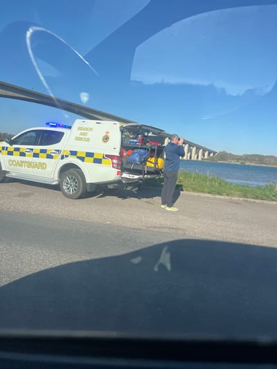 A Coastguard search and rescue vehicle is parked on a road adjacent to a body of water. A man stands at the rear of the vehicle, holding binoculars to his eyes. The vehicle is white with blue and yellow checkered markings and the words "COASTGUARD" and "SEARCH AND RESCUE" are clearly visible. The back of the vehicle is open, revealing equipment inside. The scene is outdoors, with a clear sky and a body of water. The location is likely Babergh, United Kingdom, given the Coastguard marking.