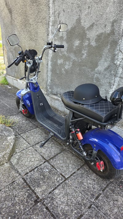 A blue electric scooter is parked outdoors, viewed from a slightly elevated, tilted perspective. The scooter features a black quilted seat with white diamond stitching, and a matte black helmet is resting on the seat. Its handlebars are chrome, fitted with two rectangular rearview mirrors. The footboard is black with a textured grip pattern, and the letters "CE" are visible on its surface. The scooter is equipped with thick tires, red brake calipers, and red suspension springs towards the rear. A kickstand supports the vehicle.

The scooter is situated on a paved surface composed of large square or rectangular tiles, each embossed with a distinct concentric circular pattern. Adjacent to the scooter is a textured concrete wall, which appears weathered. The scene is brightly lit, suggesting it is daytime. No specific activity is taking place; the scooter appears stationary and parked. The location context for this media file is Valchi Dol, Bulgaria.