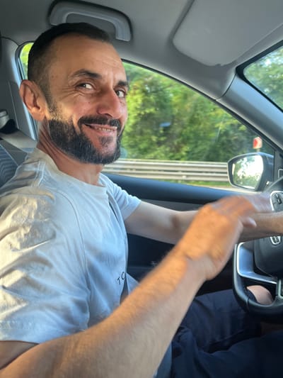 A middle-aged man with a beard and short dark hair is driving a car. He is wearing a white t-shirt and is looking at the camera with a smile. Sunlight is casting shadows on his face and arm. The car's interior is visible, including the dashboard and steering wheel. Outside the car, trees and a road are visible through the windows. The man is in Dagomys, Russia, as indicated by the context of the image.