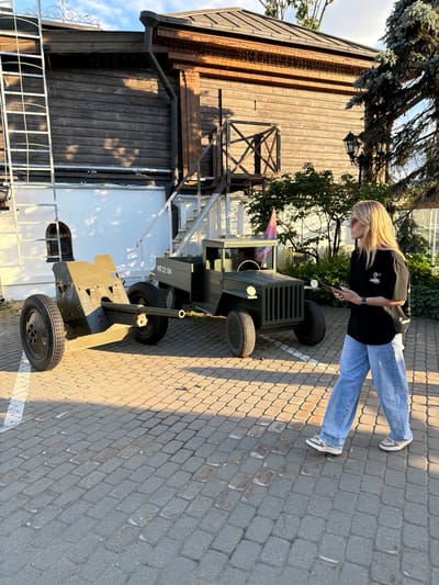 The image shows a young woman with blonde hair, wearing a black t-shirt and blue jeans, standing on a paved pathway. She is looking down at something she is holding in her hands. Beside her is a dark green, wheeled object that appears to be a replica of a historical cannon or artillery piece. The object has a barrel pointing forward and is mounted on a carriage with two wheels.
The scene is outdoors, with a wooden building visible in the background. A tree with green foliage is also present. The ground is paved with cobblestones, and shadows suggest it is daytime, likely late afternoon or early evening due to the warm light. The weather appears to be pleasant, with clear skies.
A small amount of text is visible on the wheeled object, reading "HT 21-04". The overall mood seems relaxed and casual, with the woman engaged with the historical artifact. The context provided indicates the location is Suzdal, Russia, which is known for its historical architecture and preservation sites.