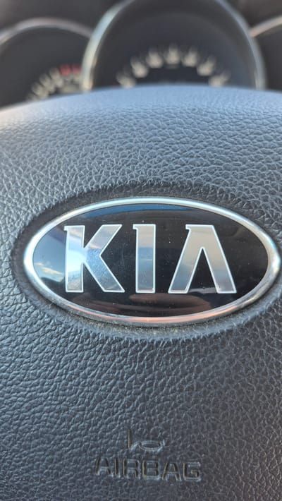 The image shows a close-up, vertically oriented shot of a car's steering wheel. The dominant subject is the silver "KIA" logo, encased in an oval, black emblem with a chrome trim. The emblem is centrally placed on the black, textured leather of the steering wheel. To the right of the emblem, embossed into the leather, is the word "AIRBAG". The background on the left of the image shows a blurred view of the car's dashboard, including what appears to be a speedometer or other instrument cluster with circular markings and some red indicator lights. The lighting suggests it is daytime, with the sun casting a slight glare on the logo and the leather's texture. The overall impression is that of a vehicle interior, likely parked or in motion, with the focus on the brand identification and safety feature. The location context provided is Novoivanovskoe, Russia, but there are no visual cues in the image to confirm this specific location. No people are visible.
