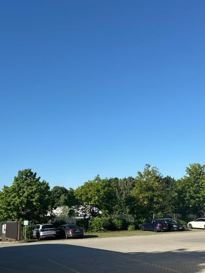 This is a vertical, eye-level, outdoor photograph taken on a clear day in Schweitenkirchen, Germany. The scene depicts a parking lot with several cars parked. The foreground is a paved parking area with yellow lines marking parking spaces. In the midground, there are at least five cars parked. On the left, a silver SUV is parked next to a dark grey sedan. Further to the right, a dark blue hatchback is parked next to a black sedan. A white car is parked to the far right. Behind the cars is a row of trees and some fencing. The background is dominated by a clear, bright blue sky. There are no people visible in the image.