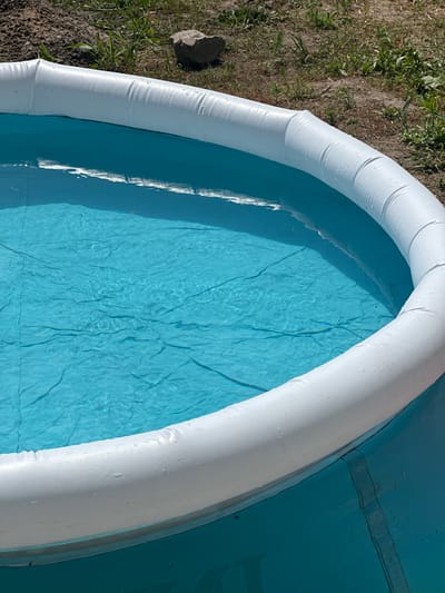 An above-ground swimming pool filled with blue water is situated outdoors on dry, grassy terrain. The pool has a white inflatable top rim. The surrounding environment shows dry soil and sparse green grass. A single gray rock is visible in the background among the grass. The scene appears to be in Yablunivka, Ukraine.