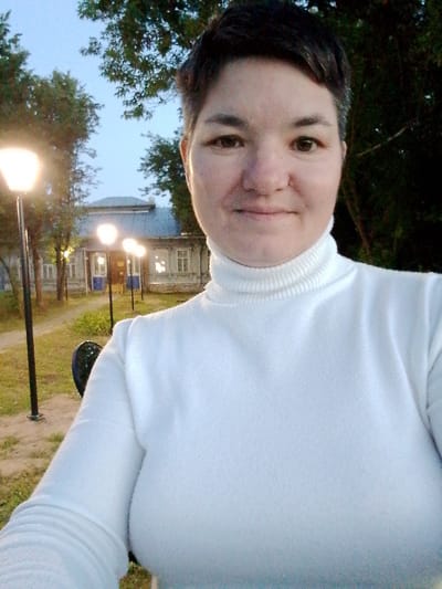 A woman is taking a selfie outdoors in Savino, Russia. She is wearing a white turtleneck sweater and has short, dark hair. The background shows a row of trees, lampposts, and a building. The sky is overcast. The lighting suggests it is either early morning or late evening.