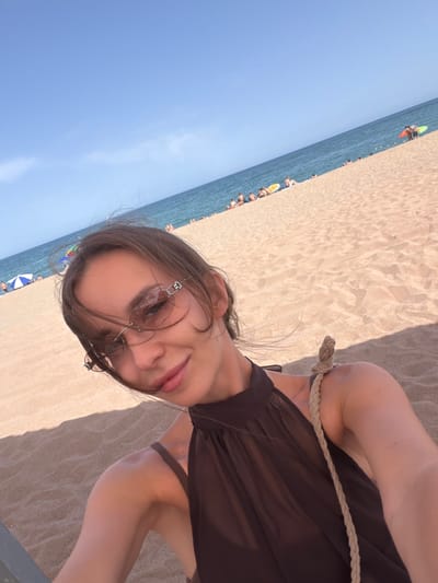 The image shows a woman on a sandy beach. She is wearing a dark brown sheer top, and her hair is dark brown and windswept. She has on sunglasses with thin, metallic frames. A rope is tied around her upper arm, and another rope is wrapped around her torso. The beach is populated with other beachgoers, some of whom are in the water or on floats. Beach umbrellas are visible in the distance. The water is blue and appears calm. The sky is clear and blue. The scene suggests a sunny day at the beach, likely during daylight hours. The overall mood seems relaxed and casual, with the woman posing for the camera. The location is Sant Adrià de Besòs, Spain.