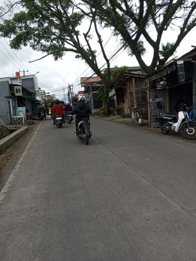 A daytime street scene in Melakudikadu, India, is depicted under an overcast sky. A paved road, showing a longitudinal seam, is the central feature. Five individuals are visible, each operating a motorcycle and traveling away from the camera. The lead motorcyclist wears a black jacket and a white full-face helmet. Behind them, a rider in a red jacket is followed by another wearing a light pink helmet. Two additional motorcyclists are visible further back in the procession.
On the left side of the road, a concrete curb separates the asphalt from a row of structures. These buildings, appearing to be a mix of residential and commercial, feature varied facades, utility meters, and signage, including a partially visible sign with green text and a red water tank on one rooftop. Multiple power lines and utility poles are strung overhead along this side of the street.
On the right side of the road, a large tree with a dense, gnarled, moss-covered canopy extends over the thoroughfare. Below the tree, a rustic wooden stall with displayed items and a corrugated metal structure are present. A light-colored bicycle is parked near the wooden stall. Further right, a white and black motorcycle is parked, facing the camera, with a dark-colored motorcycle partially visible behind it. Distant hills or mountains are discernible beneath the cloudy horizon in the background.